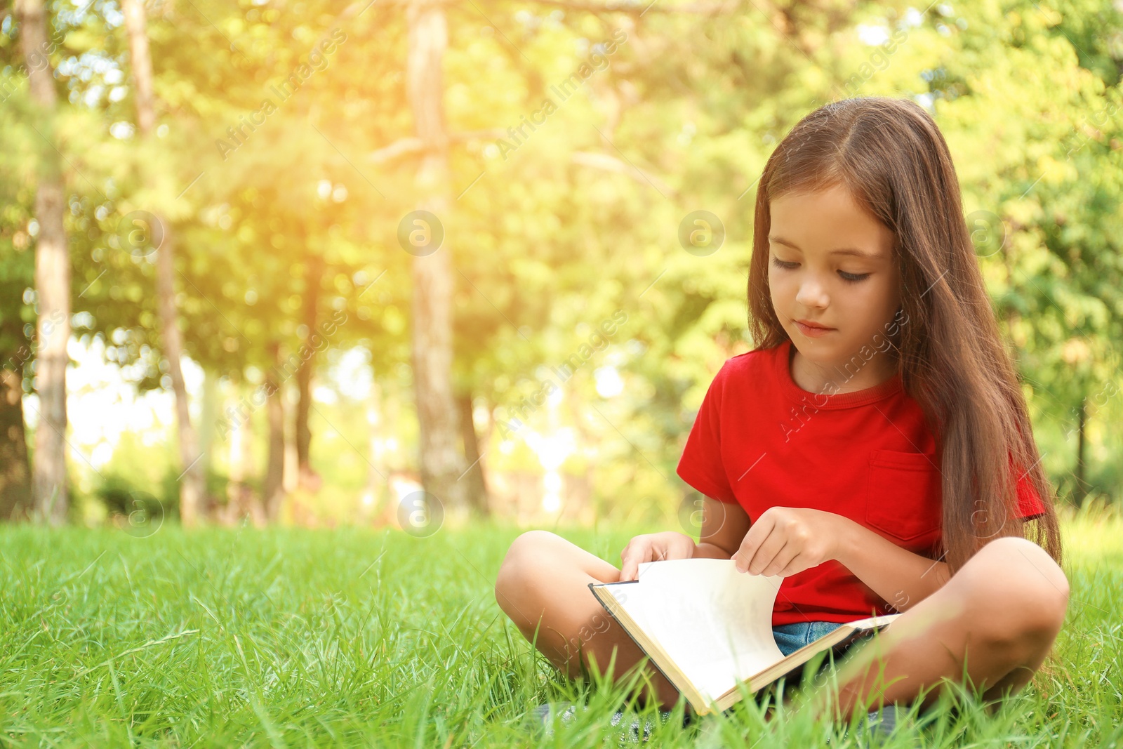 Photo of Cute little girl reading book on green grass in park