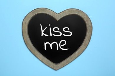 Photo of Small heart shaped chalkboard with phrase Kiss Me on light blue background, top view