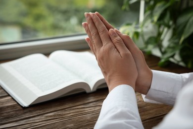 Photo of Woman holding hands clasped while praying at wooden table with Bible, closeup. Space for text