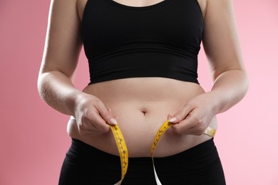 Photo of Woman measuring belly with tape on pink background, closeup. Overweight problem