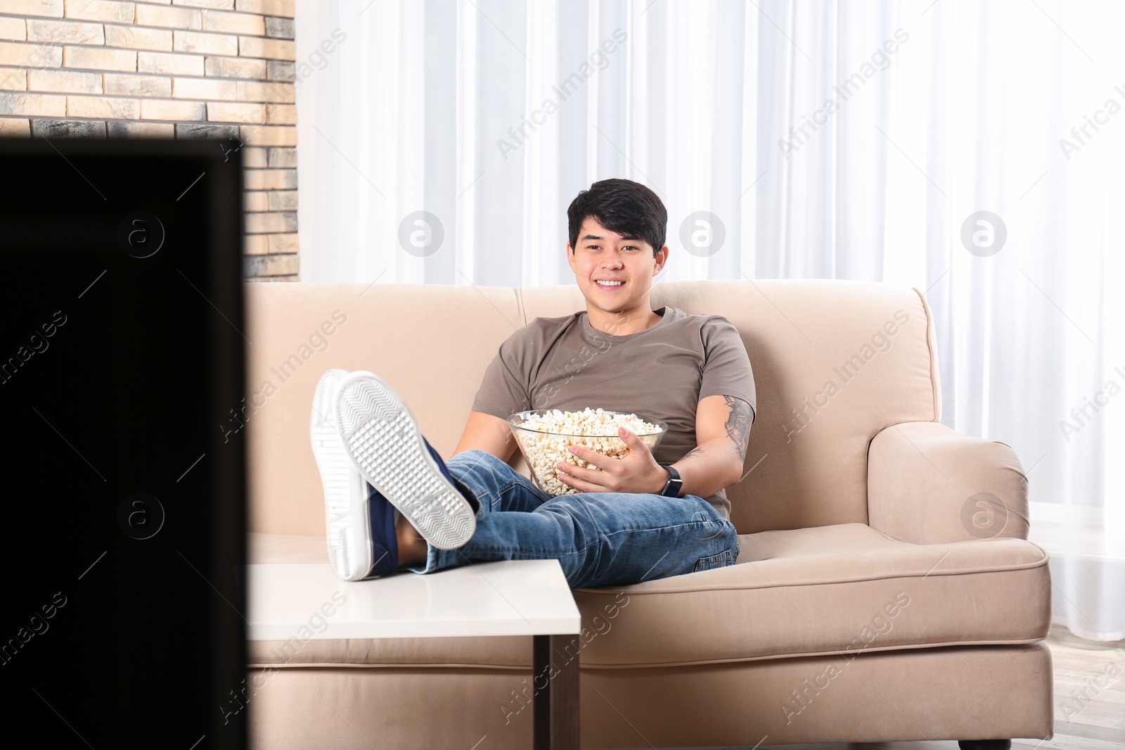 Photo of Young man with bowl of popcorn watching TV on sofa at home