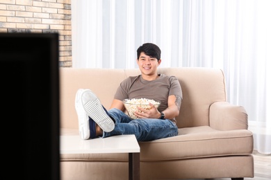 Photo of Young man with bowl of popcorn watching TV on sofa at home