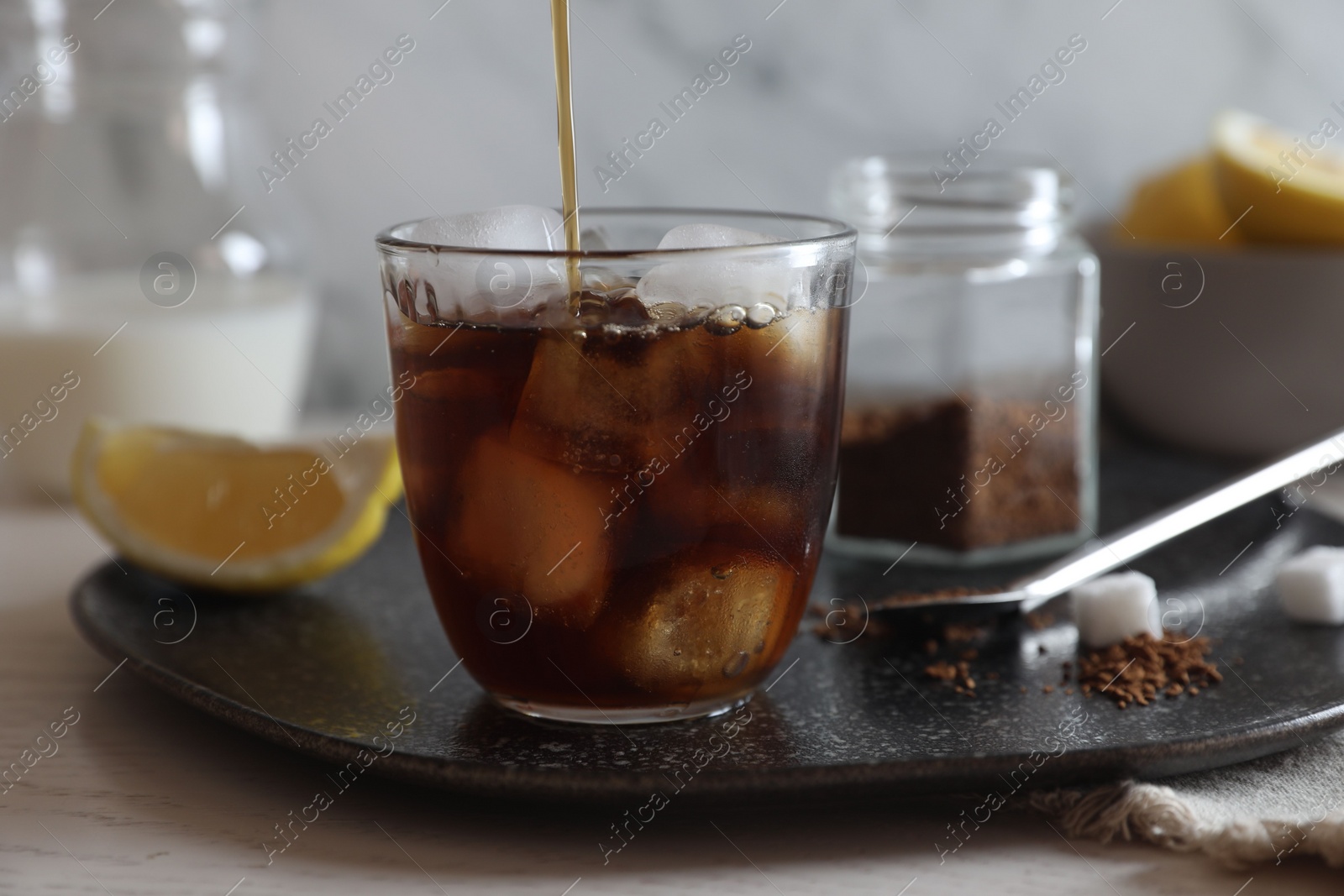 Photo of Pouring coffee into glass with ice cubes at white wooden table, closeup