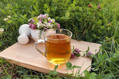 Photo of Cup of aromatic herbal tea, pestle and ceramic mortar with different wildflowers on wooden board in meadow