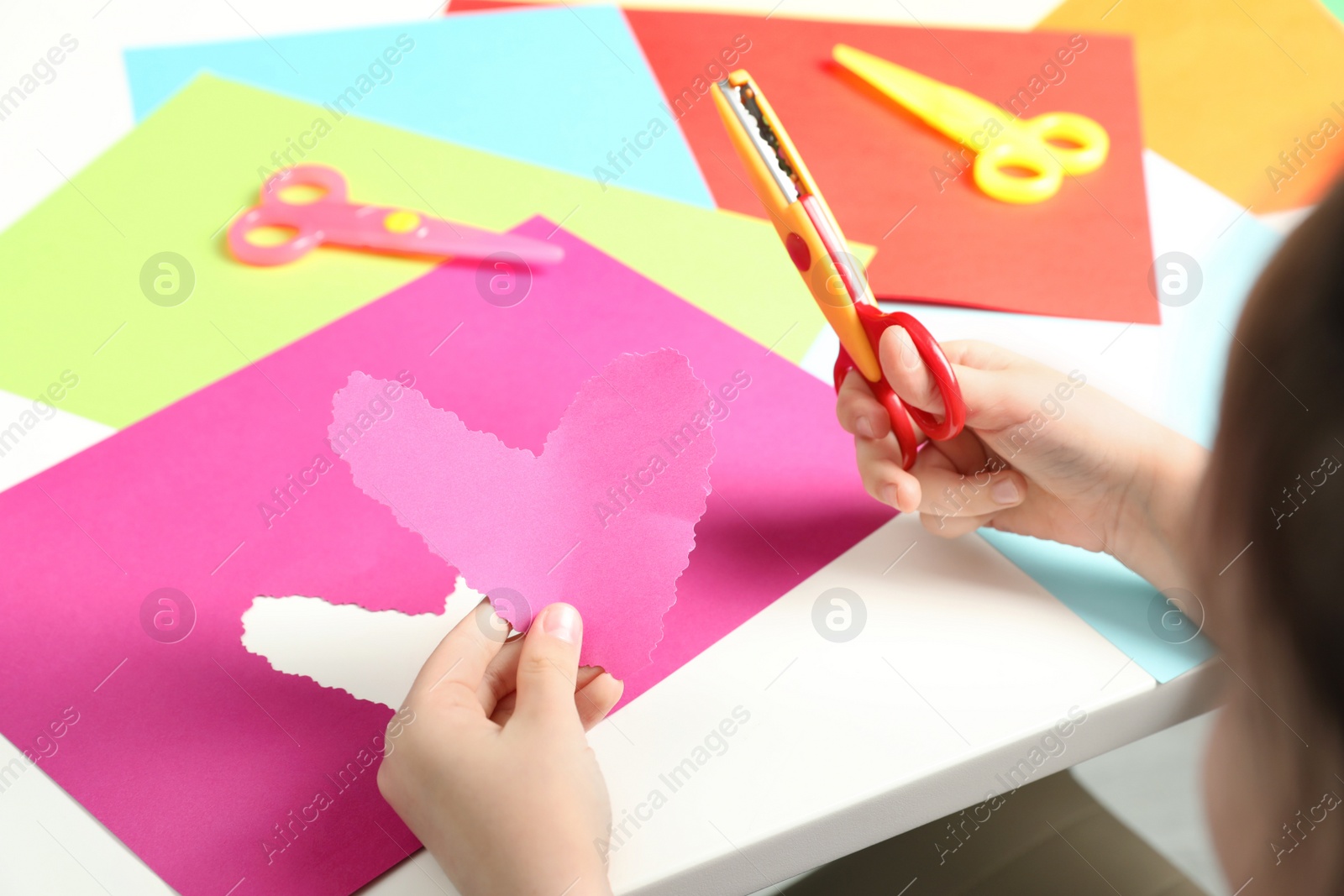 Photo of Child cutting out paper heart with craft scissors at table, closeup