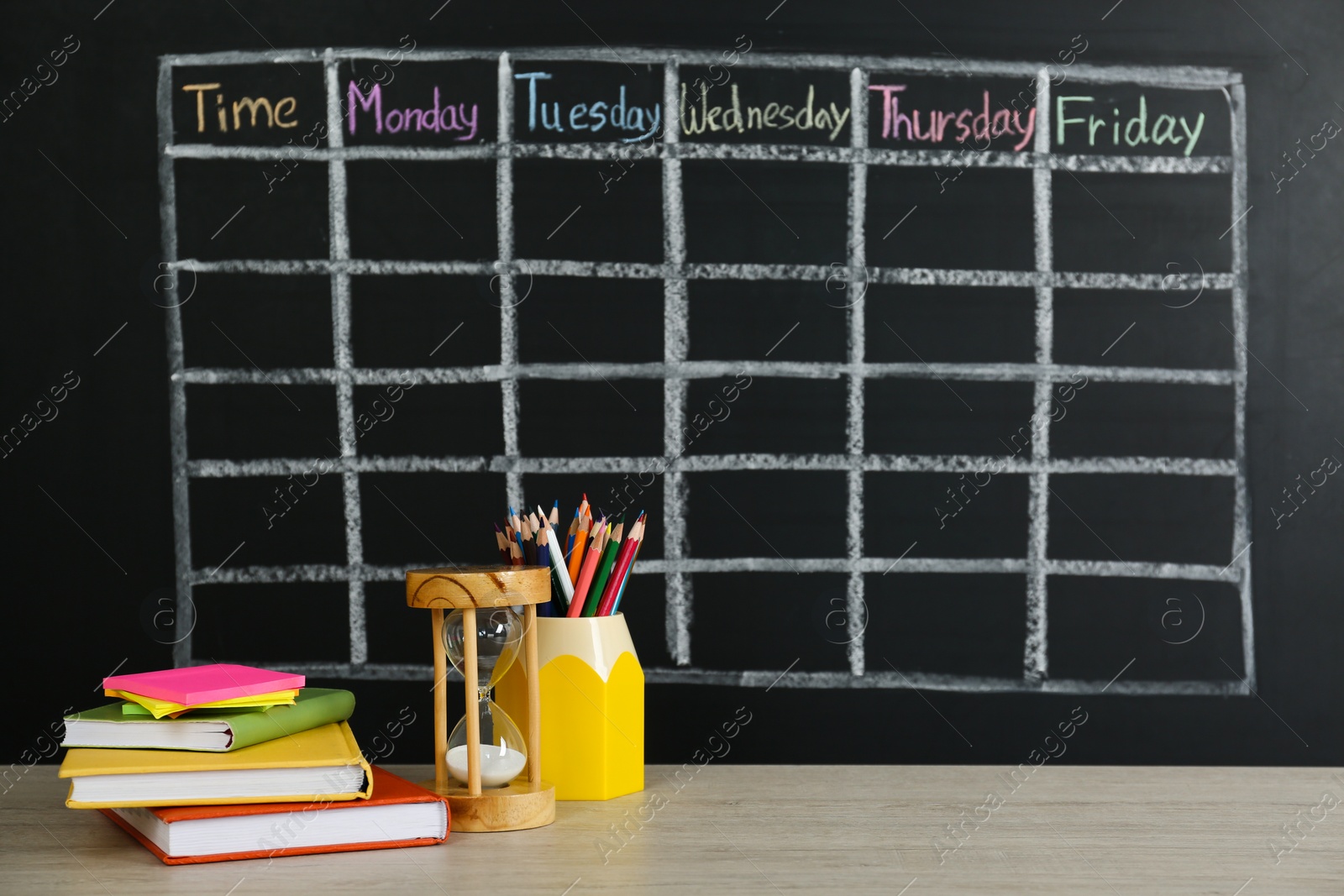 Photo of Hourglass and stationery on white wooden table near blackboard with drawn school timetable