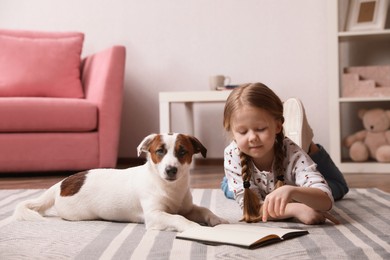 Photo of Cute little girl with her dog reading book on carpet at home. Childhood pet