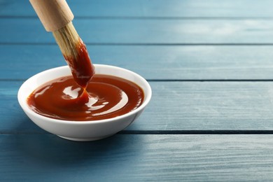 Tasty barbeque sauce in bowl and brush on blue wooden table, closeup. Space for text