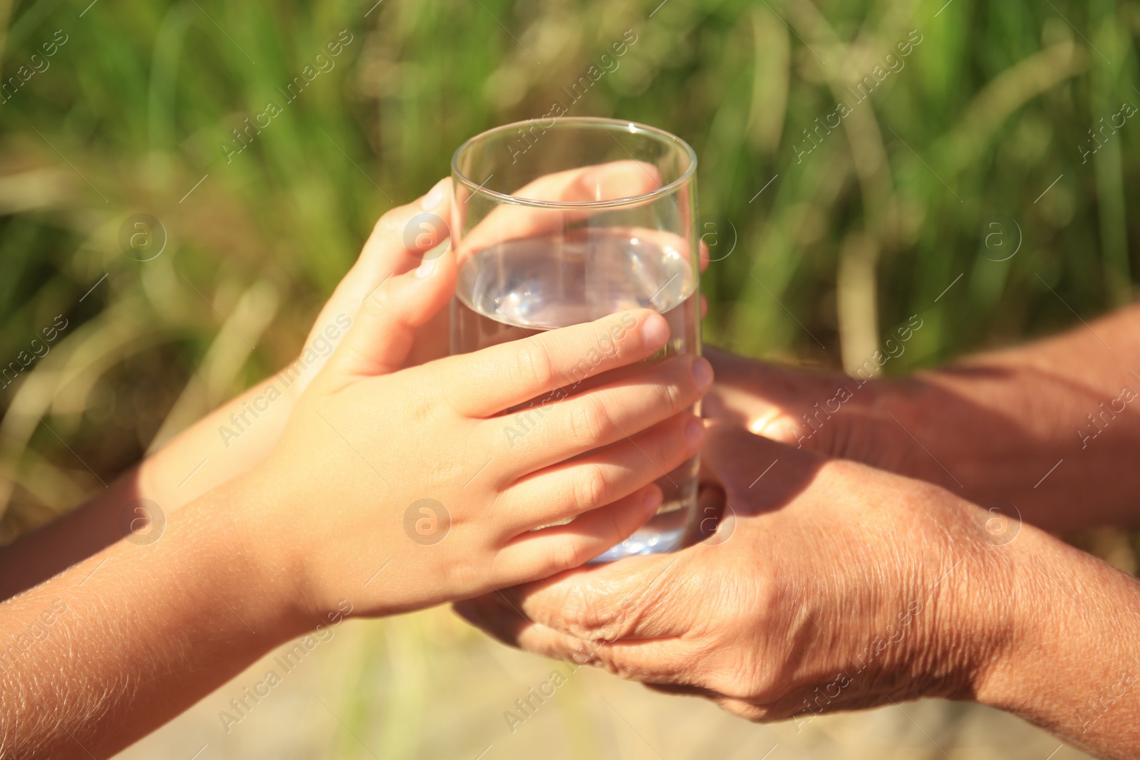 Photo of Child giving glass of water to elderly woman outdoors on sunny day, closeup