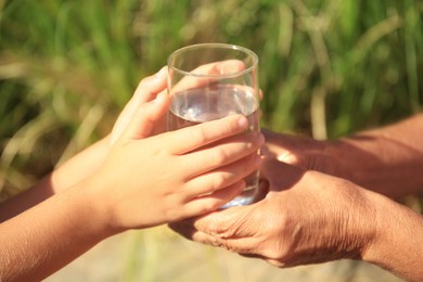 Photo of Child giving glass of water to elderly woman outdoors on sunny day, closeup