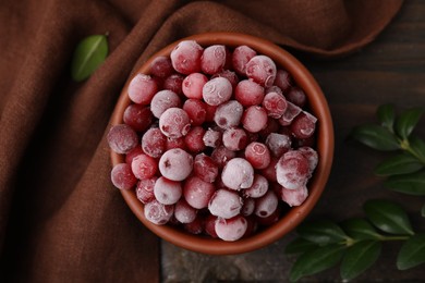 Frozen red cranberries in bowl and green leaves on wooden table, top view