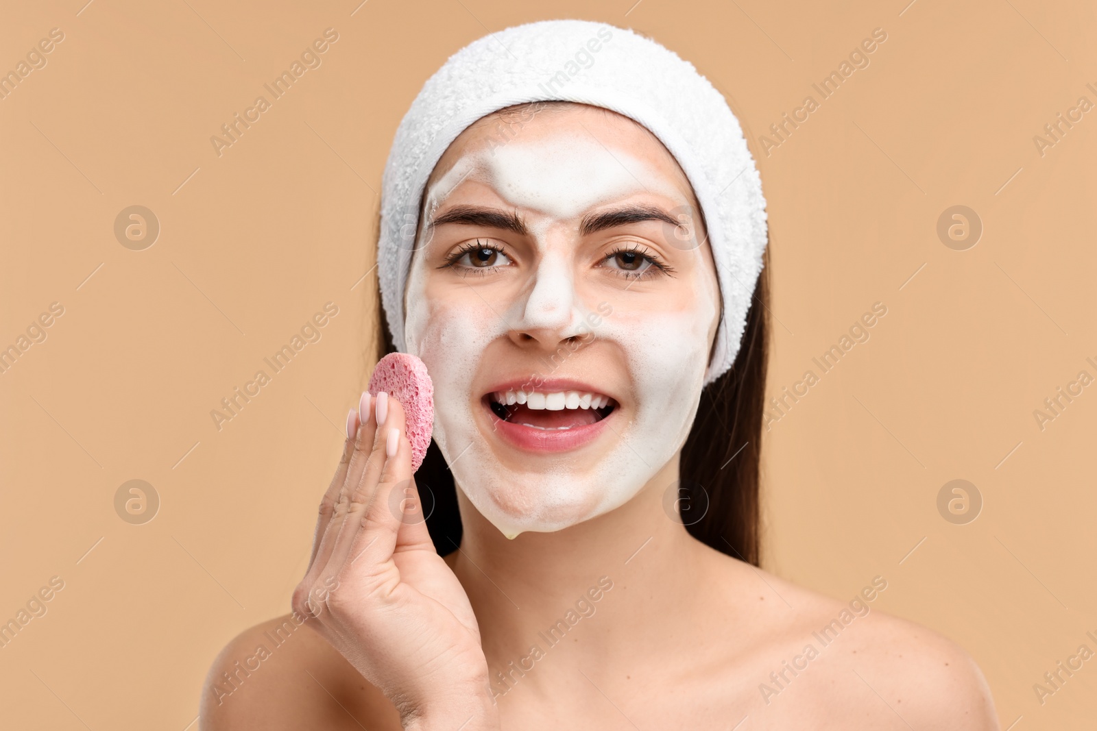 Photo of Young woman with headband washing her face using sponge on beige background