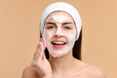 Young woman with headband washing her face using sponge on beige background