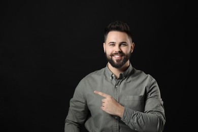 Young man with mustache pointing at something on black background. Space for text