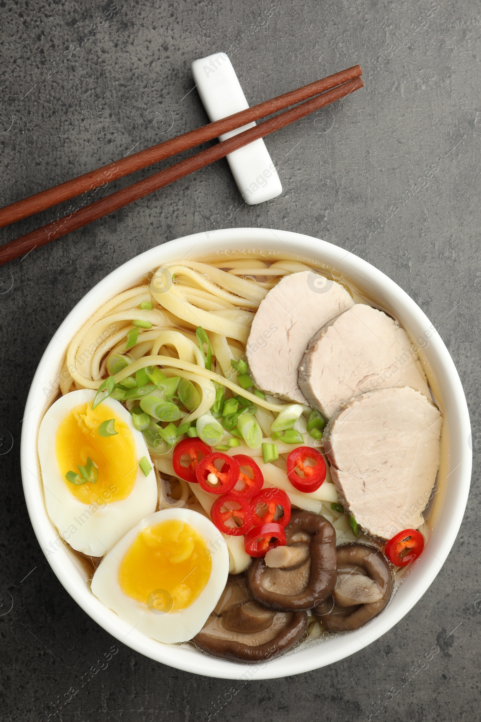 Photo of Delicious ramen with meat in bowl and chopsticks on grey textured table, top view. Noodle soup