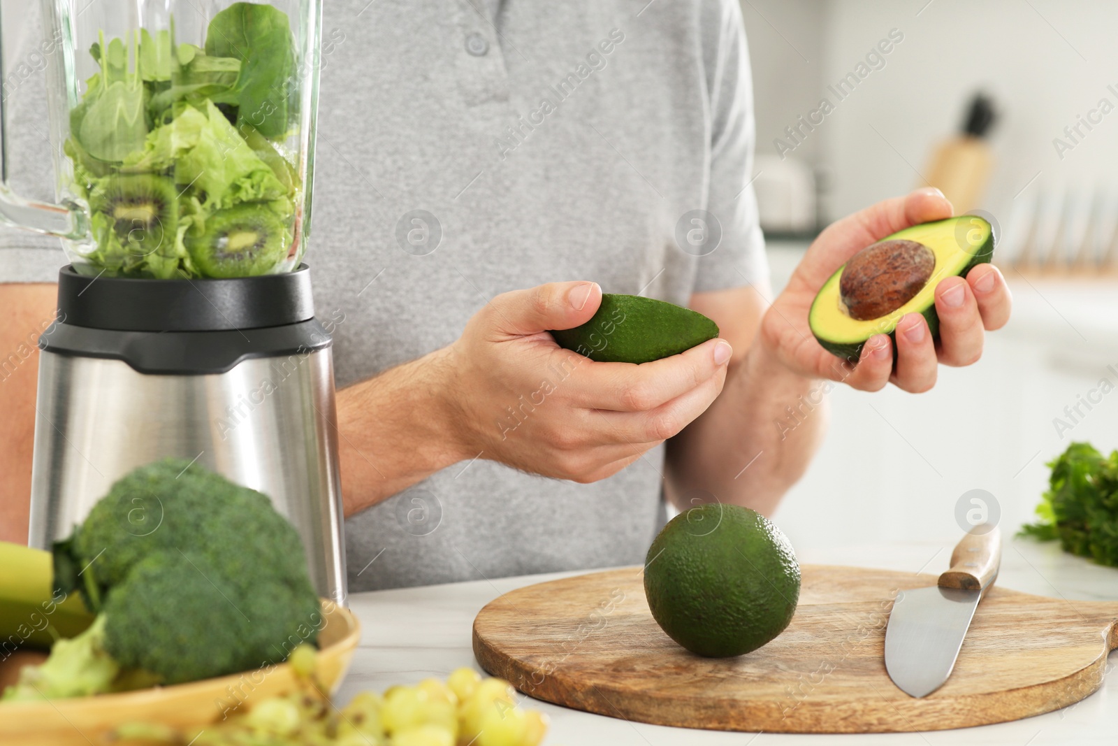 Photo of Man holding halves of avocado for delicious smoothie at white table in kitchen, closeup