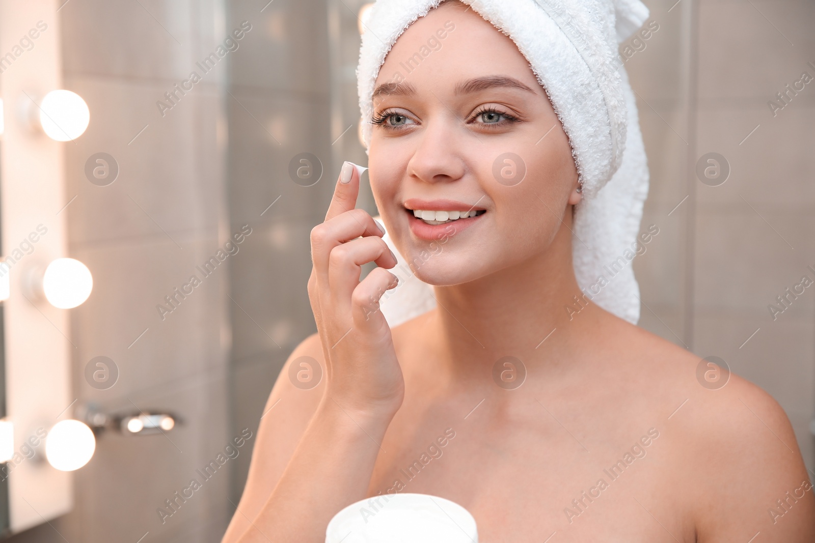 Photo of Beautiful woman with towel on head applying face cream in bathroom