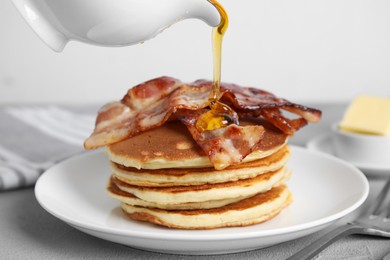 Photo of Pouring maple syrup onto delicious pancakes with fried bacon at grey table, closeup