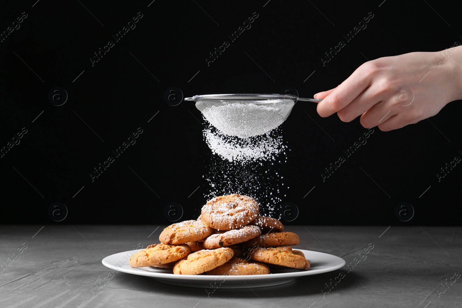 Photo of Woman with sieve sprinkling powdered sugar onto cookies at grey textured table, closeup