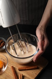 Photo of Woman whipping ingredients with mixer at black table, closeup. Cooking delicious eggnog