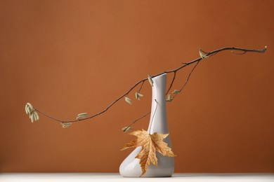Vase with tree branch and autumn leaf on white table against brown background