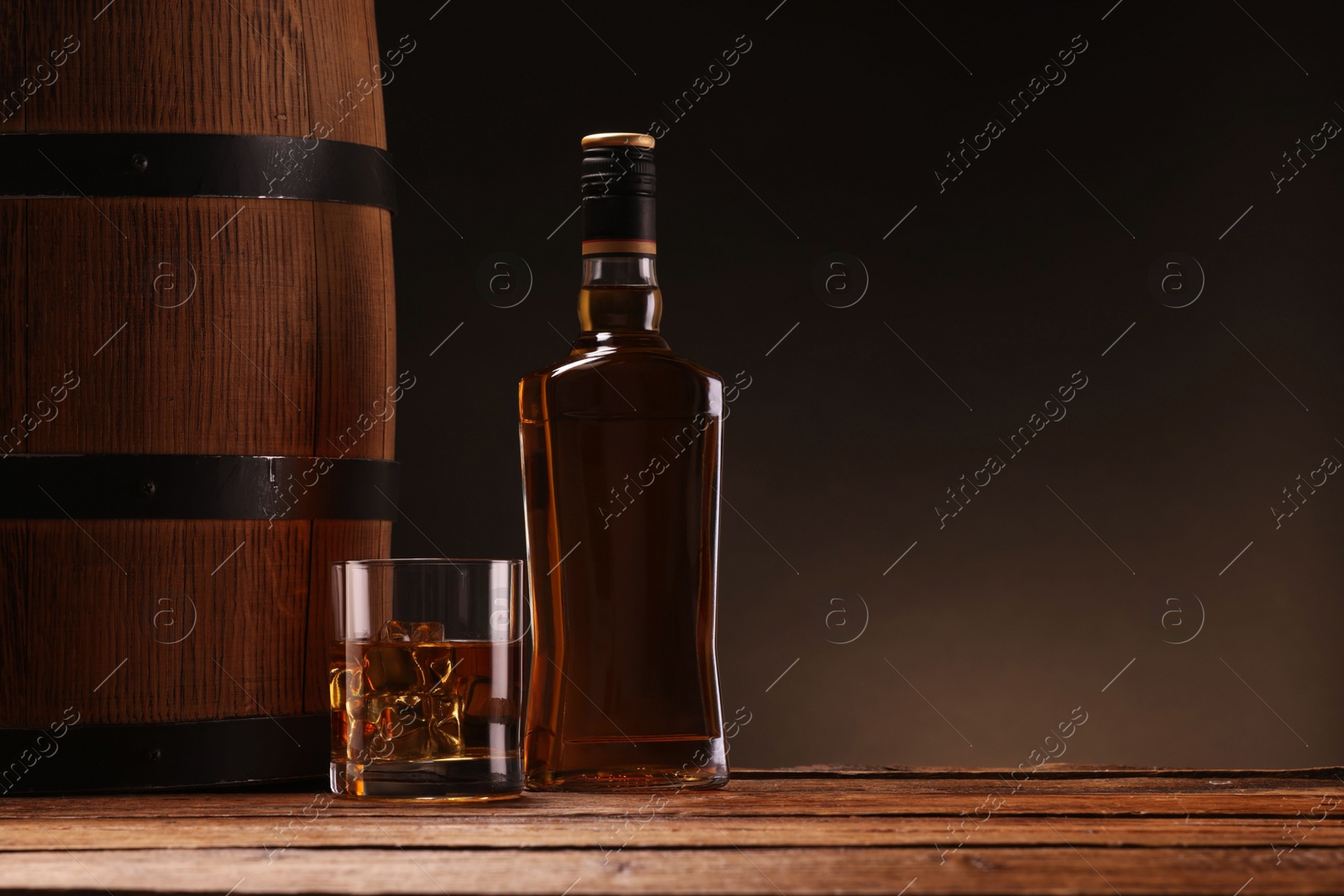 Photo of Whiskey with ice cubes in glass, bottle and barrel on wooden table against black background, space for text