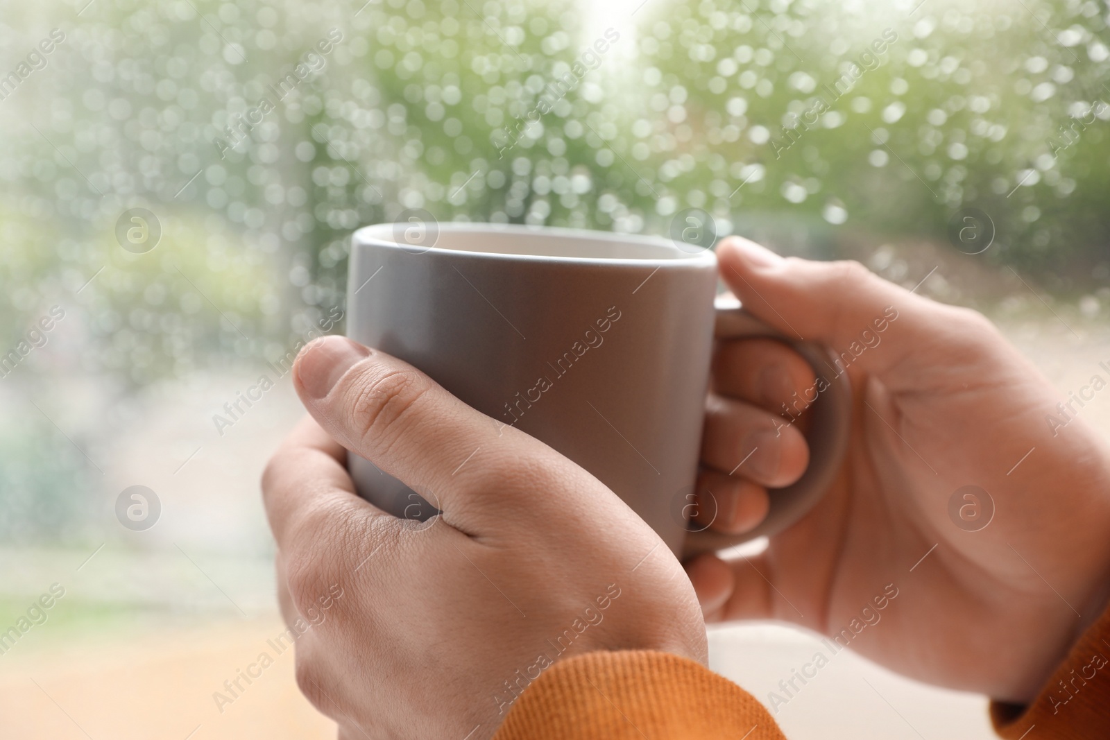 Photo of Young man with cup of coffee near window indoors on rainy day, closeup