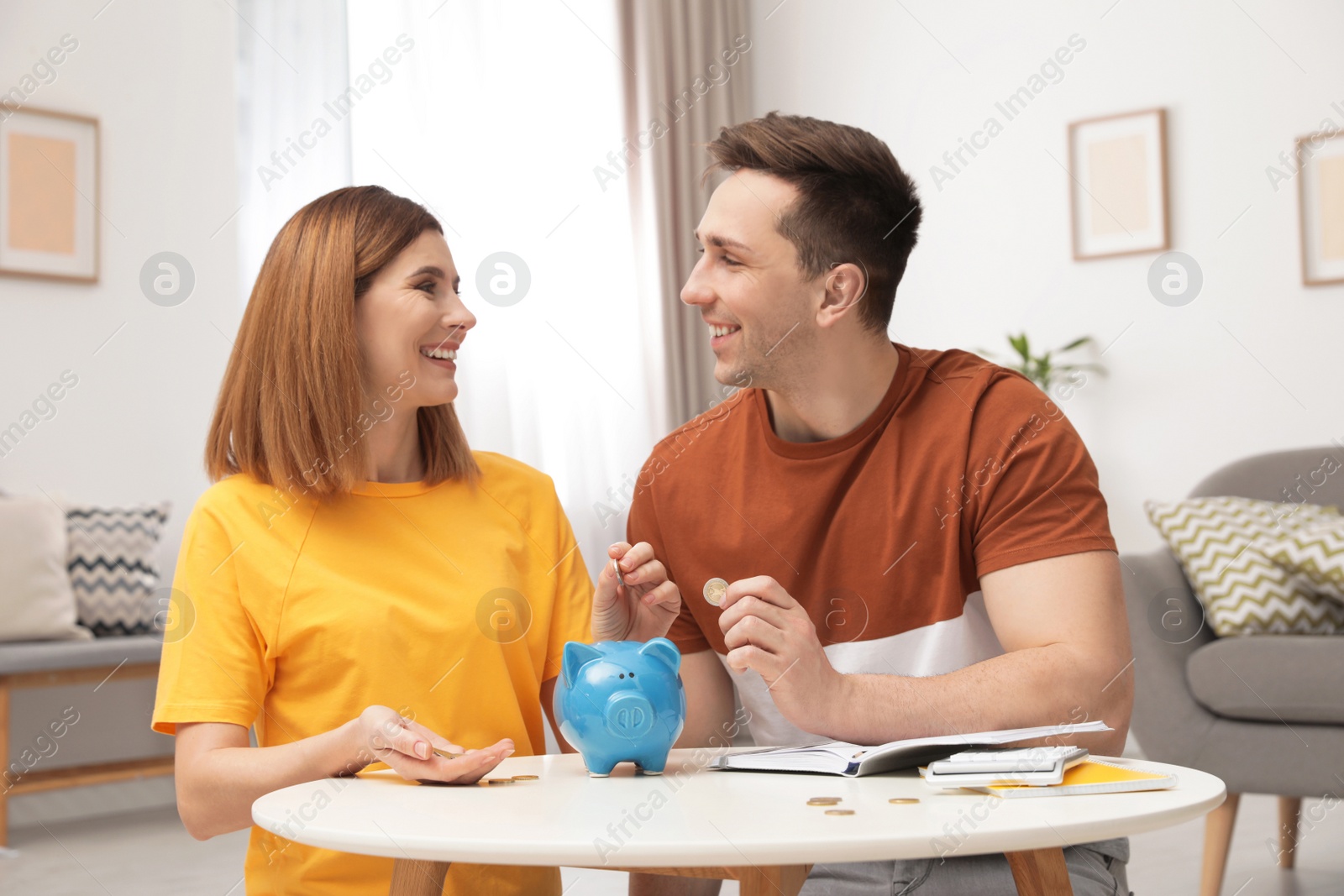 Photo of Couple putting coins into piggy bank at table in living room. Saving money