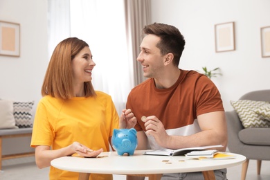 Photo of Couple putting coins into piggy bank at table in living room. Saving money