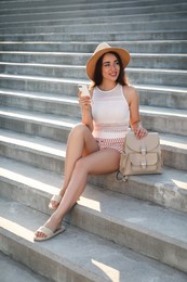 Photo of Beautiful young woman with stylish backpack and smartphone sitting on stairs outdoors
