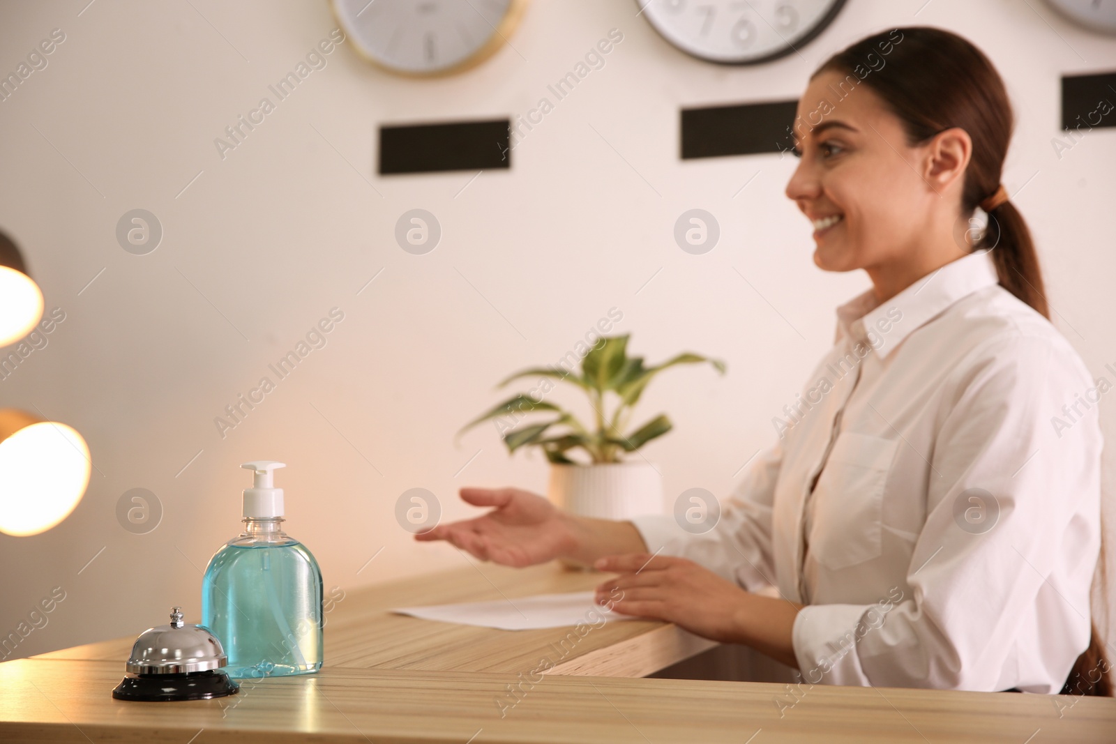 Photo of Receptionist at countertop in hotel, focus on dispenser bottle with antiseptic gel and service bell