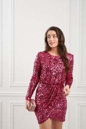 Beautiful young woman with clutch in stylish pink sequin dress near white wall indoors. Party outfit