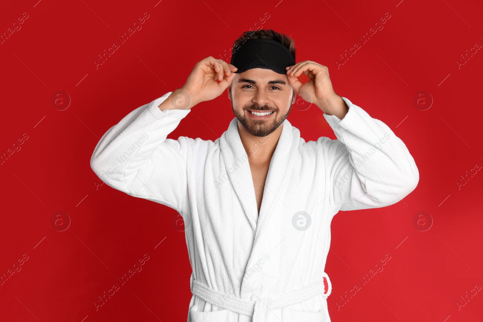 Photo of Happy young man in bathrobe and eye sleeping mask on red background