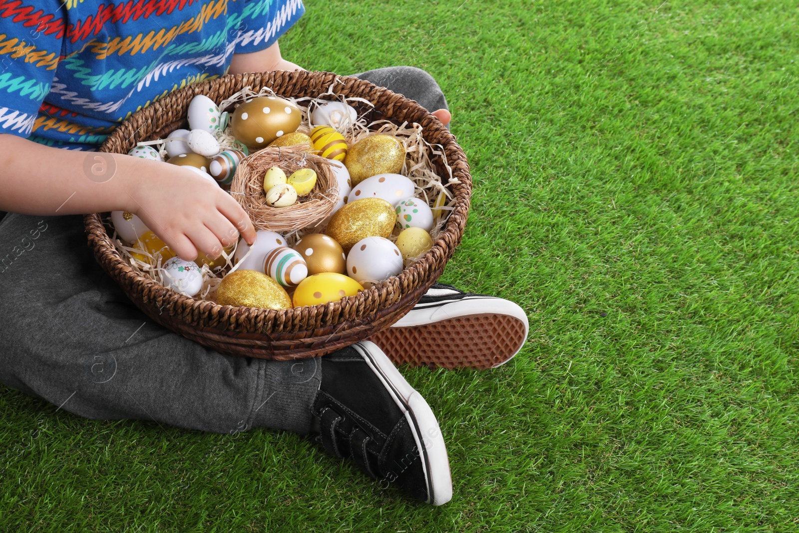 Photo of Little boy with basket full of dyed Easter eggs on green grass, closeup