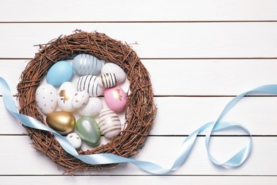 Photo of Festively decorated Easter eggs, vine wreath and light blue ribbon on white wooden table, flat lay
