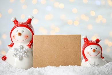 Photo of Decorative snowmen near blank greeting card on artificial snow against blurred festive lights, space for text