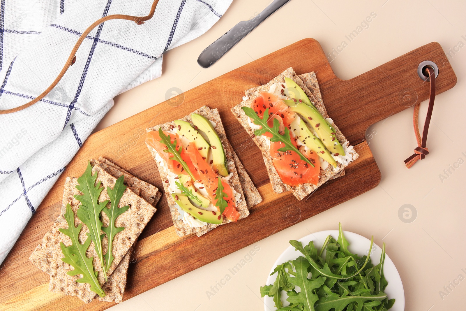 Photo of Fresh crunchy crispbreads with cream cheese, salmon, avocado and arugula on beige table, flat lay