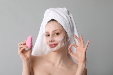 Young woman washing face with brush and cleansing foam on grey background