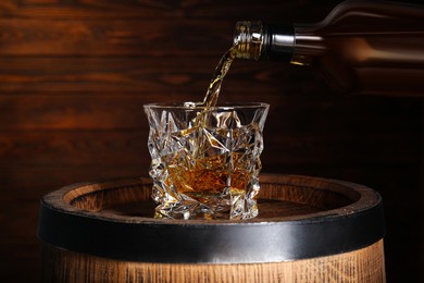 Photo of Pouring whiskey into glass from bottle on wooden barrel against brown background
