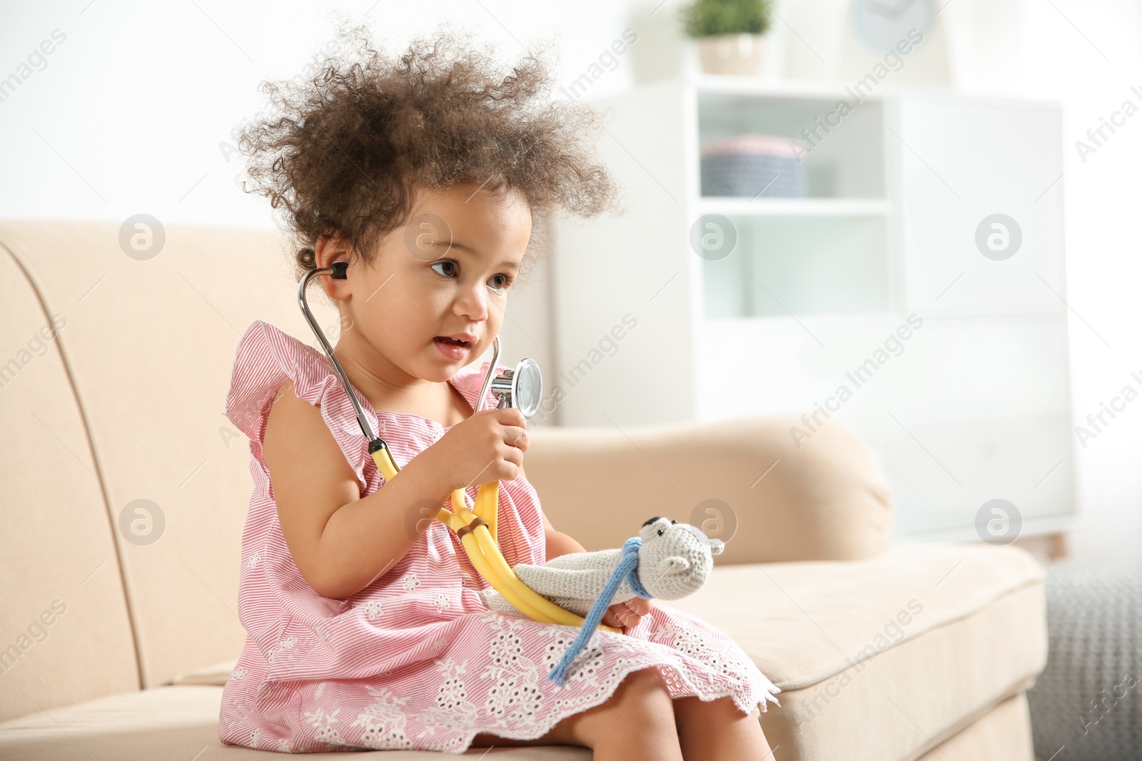 Photo of Cute African American child imagining herself as doctor while playing with stethoscope and toy on couch at home