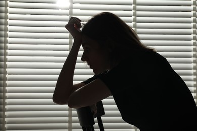 Photo of Silhouette of sad young woman near closed blinds indoors