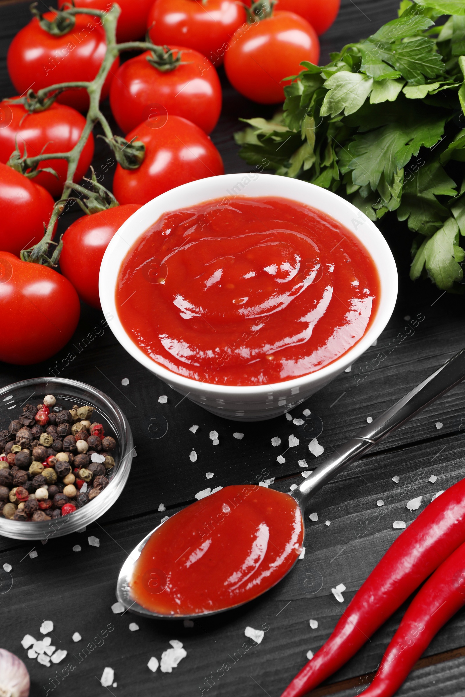 Photo of Delicious ketchup in bowl, spices and products on black wooden table. Tomato sauce