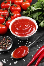 Photo of Delicious ketchup in bowl, spices and products on black wooden table. Tomato sauce