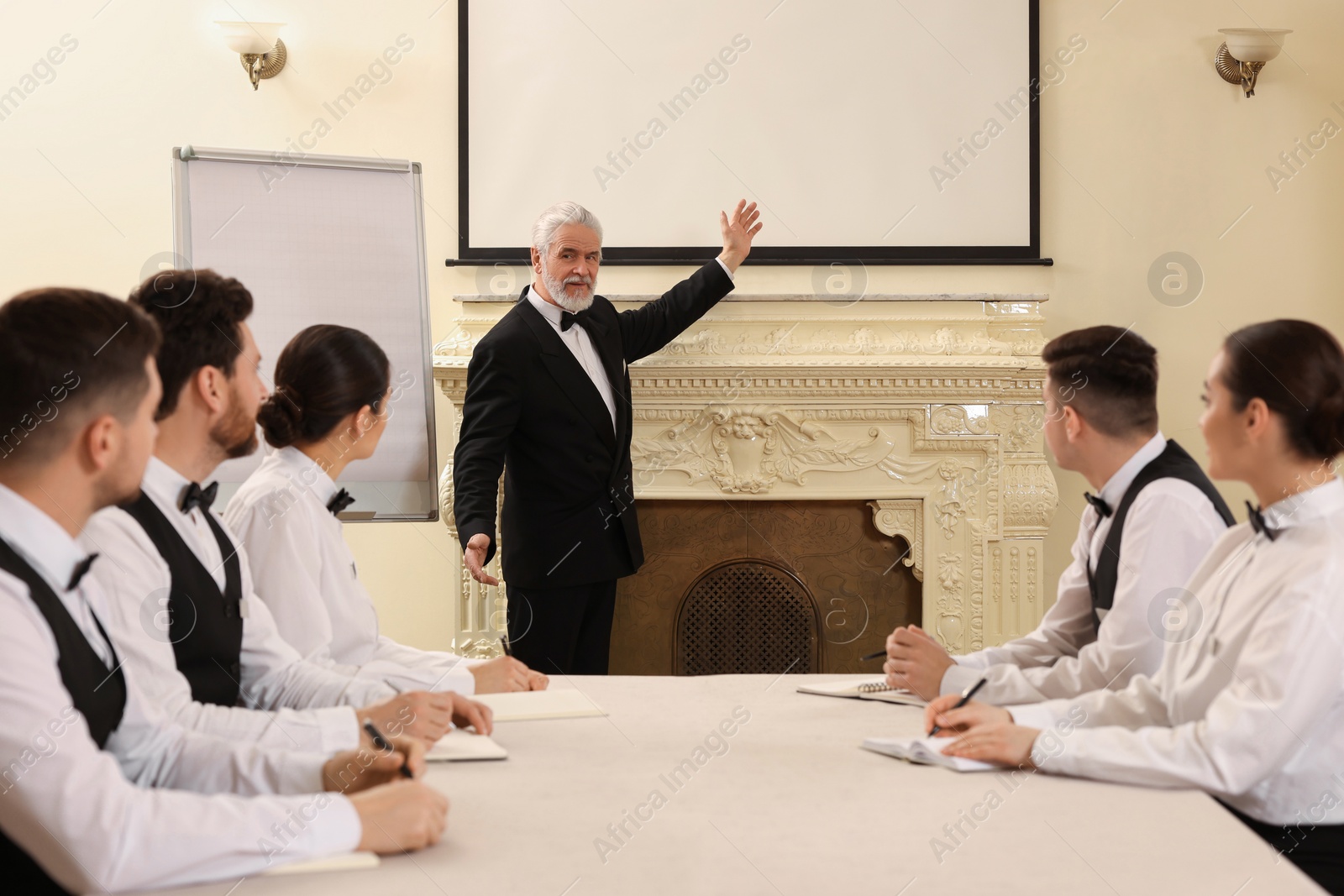 Photo of Senior man in formal suit teaching trainees indoors. Professional butler courses