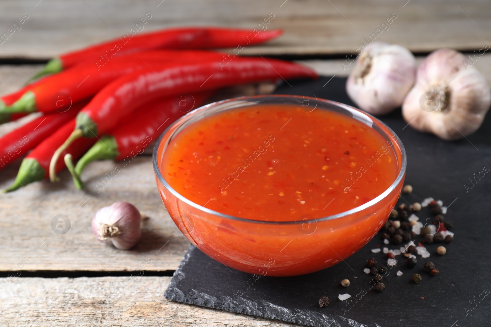 Photo of Spicy chili sauce in bowl and ingredients on wooden table, closeup