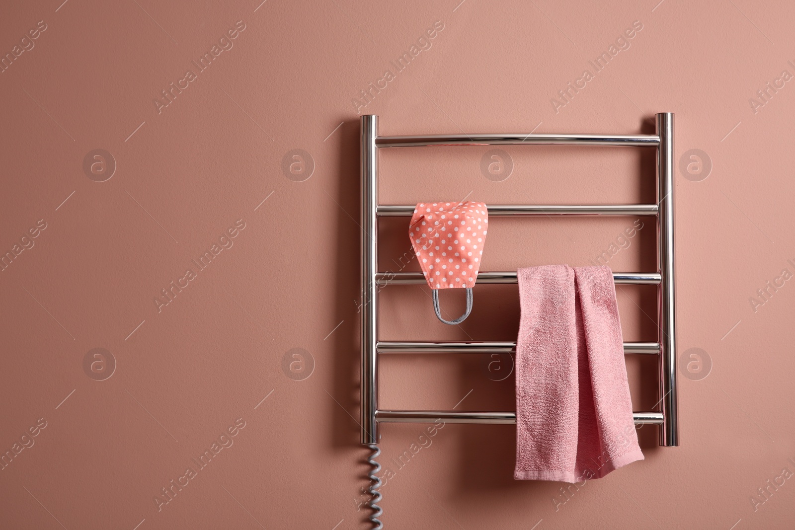 Photo of Modern heated towel rail with cloth face mask and towel on light pink wall. Space for text