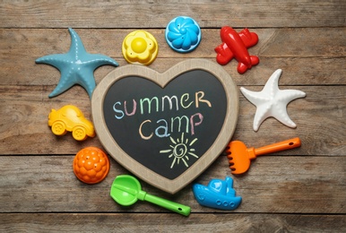 Photo of Heart shaped chalkboard with text SUMMER CAMP and different sand molds on wooden background, flat lay