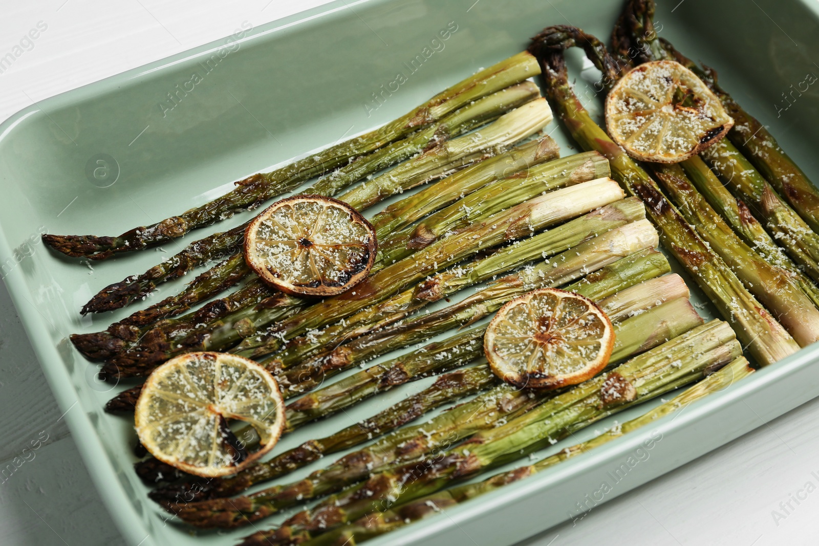 Photo of Oven baked asparagus with lemon slices in ceramic dish on white table, closeup