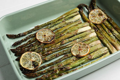 Photo of Oven baked asparagus with lemon slices in ceramic dish on white table, closeup