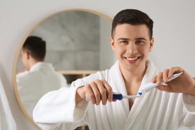 Photo of Happy man squeezing toothpaste from tube onto electric toothbrush in bathroom